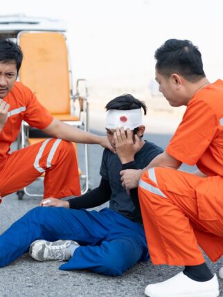 Asian male EMS paramedics or emergency medical technicians in orange uniforms performing first aid to patient with leg injury from road accident